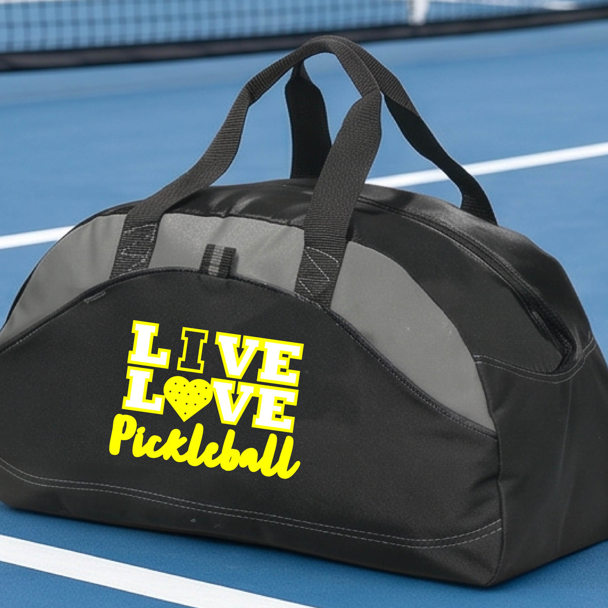 A black and gray duffel bag with yellow text reading 'LIVE LOVE Pickleball' sits on a blue pickleball court. The bag has dual top handles and a front zipper pocket.