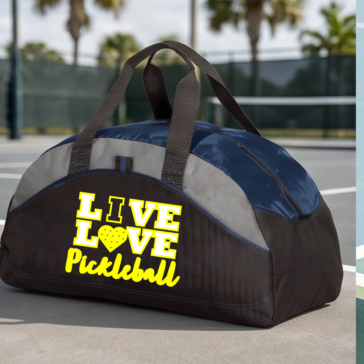 A red, black, and gray duffel bag with yellow text reading 'LIVE LOVE Pickleball' sits on a pickleball court. The bag features black handles, a red and gray upper panel, and a black lower section with red piping.