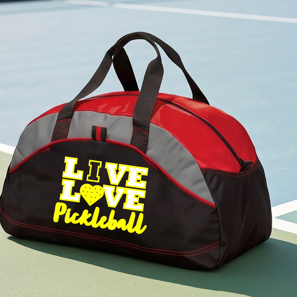 A red, black, and gray duffel bag with yellow text reading 'LIVE LOVE Pickleball' sits on a pickleball court. The bag features black handles, a red and gray upper panel, and a black lower section with red piping.