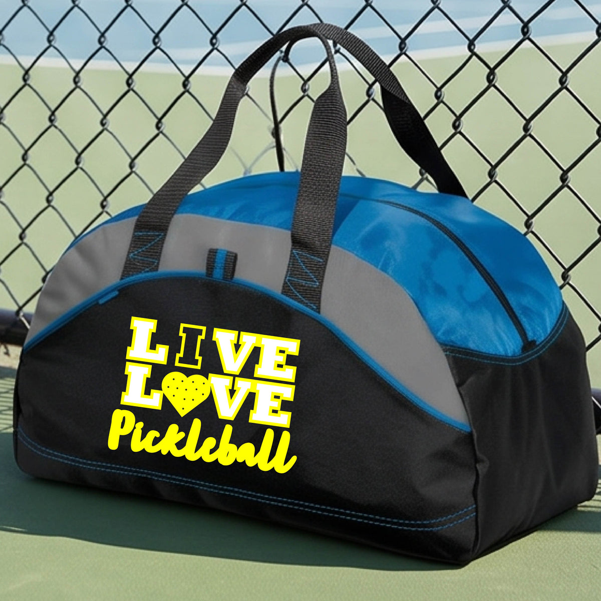 A black and blue pickleball duffel bag with gray panels and yellow 'LIVE LOVE Pickleball' text, placed on a green pickleball court in front of a chain-link fence.