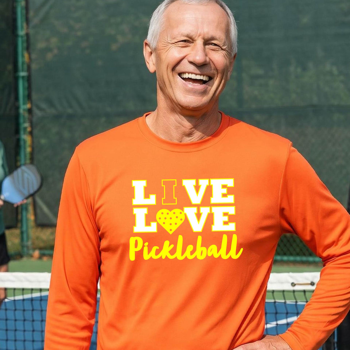 A man on a pickleball court wearing an orange long-sleeve V-neck shirt with a yellow graphic that reads 'LIVE LOVE Pickleball' and a heart-shaped pickleball dot pattern inside the word 'LOVE'.