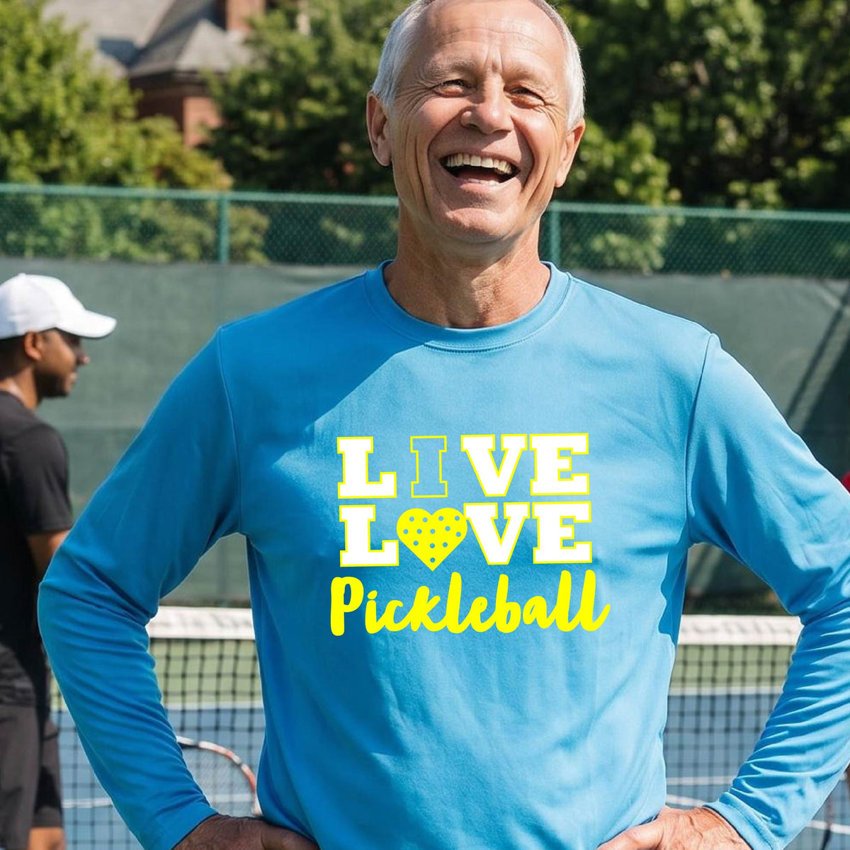 A man on a pickleball court wearing a bright blue long-sleeve V-neck shirt with a yellow graphic that reads 'LIVE LOVE Pickleball' and a heart-shaped pickleball dot pattern inside the word 'LOVE'.