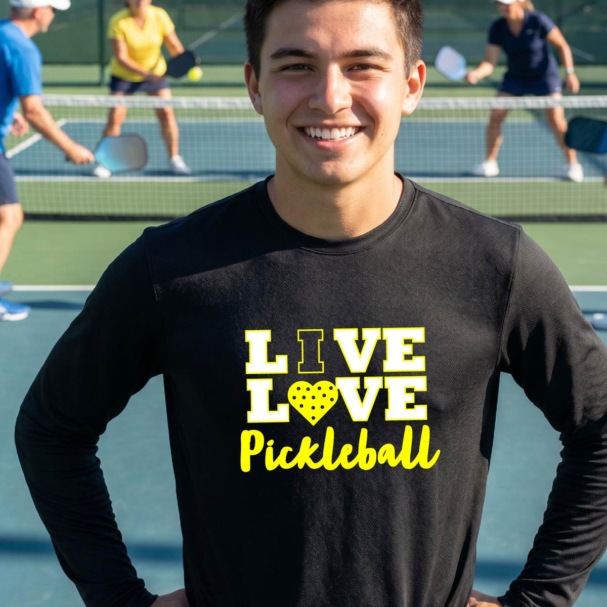 A man on a pickleball court wearing a black long-sleeve V-neck shirt with a yellow graphic that reads 'LIVE LOVE Pickleball' and a heart-shaped pickleball dot pattern inside the word 'LOVE'.