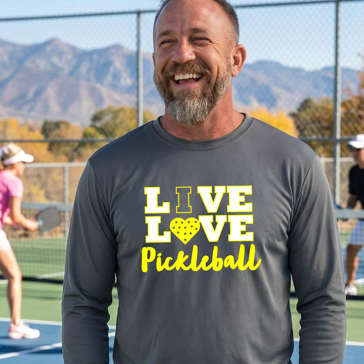 A man on a pickleball court wearing a gray long-sleeve V-neck shirt with a yellow graphic that reads 'LIVE LOVE Pickleball' and a heart-shaped pickleball dot pattern inside the word 'LOVE'.