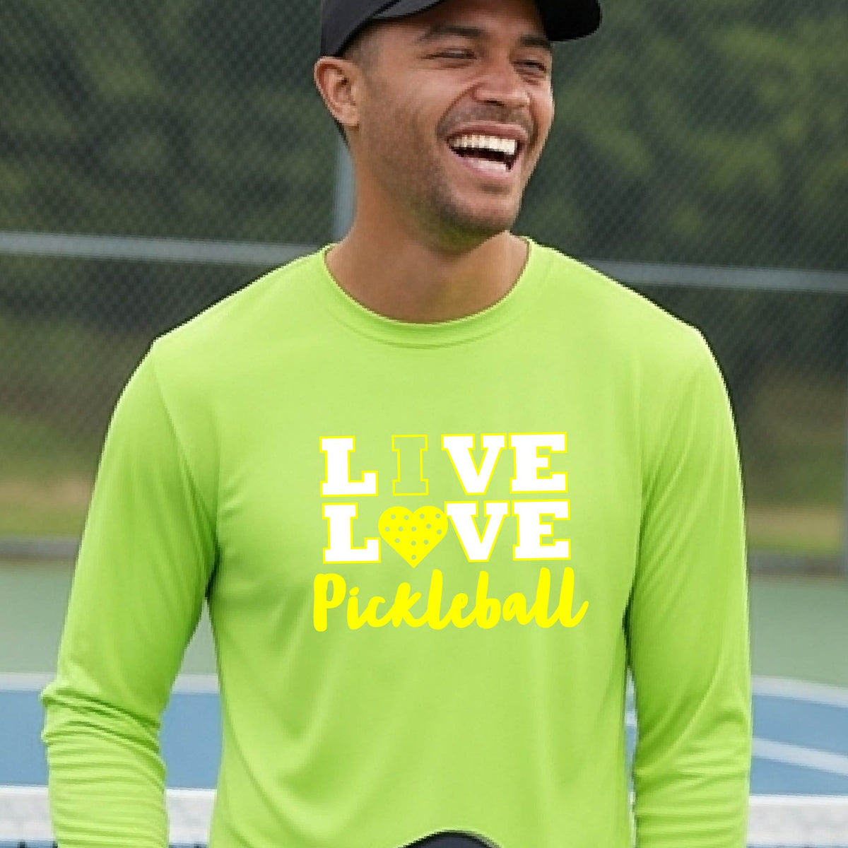 A man on a pickleball court wearing a lime green long-sleeve V-neck shirt with a yellow graphic that reads 'LIVE LOVE Pickleball' and a heart-shaped pickleball dot pattern inside the word 'LOVE'.