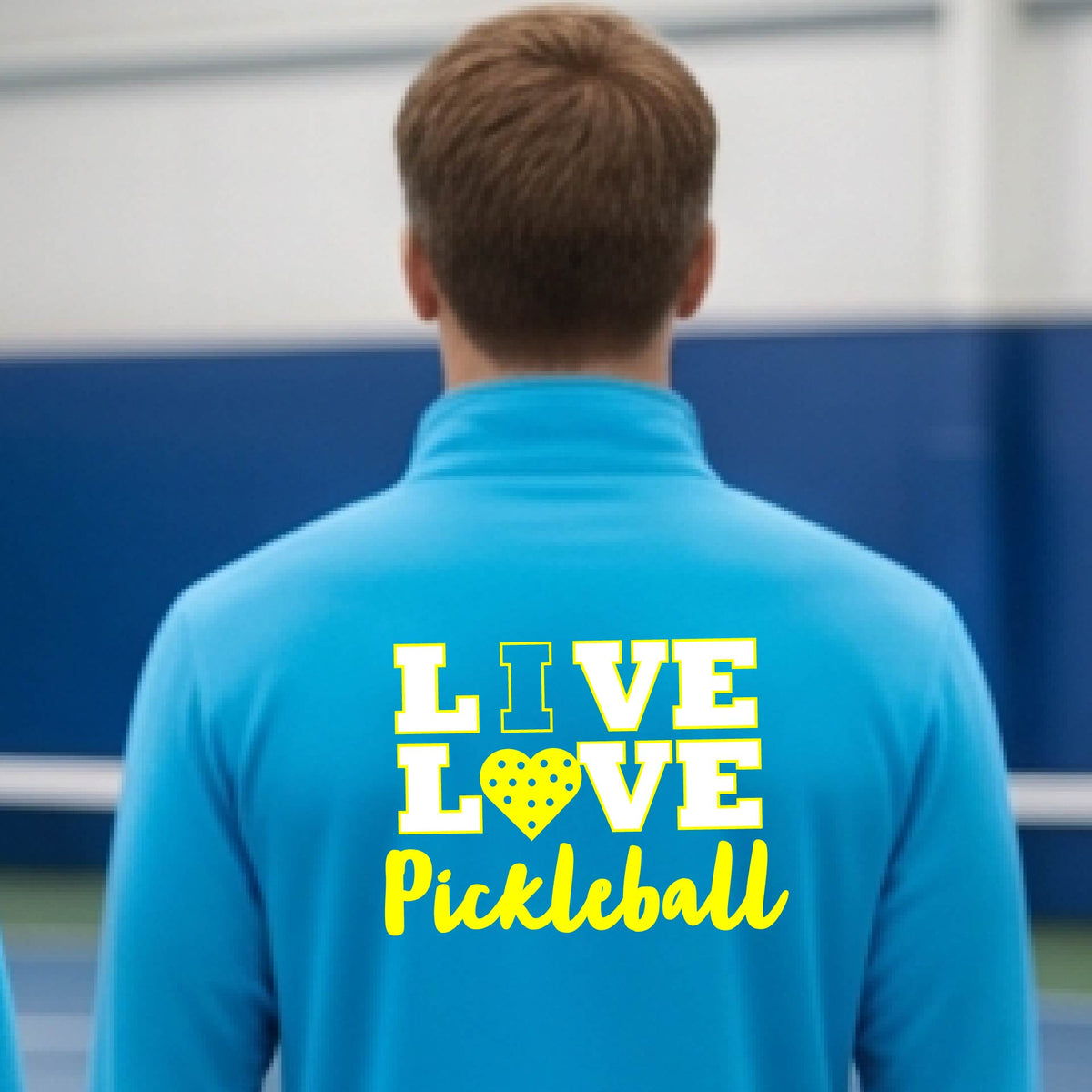 A man viewed from behind wearing a bright blue long-sleeve athletic quarter-zip shirt with a yellow graphic on the back that reads 'LIVE LOVE Pickleball'  with a pickleball motiff heart for replacing the O,set against an outdoor blurred background.