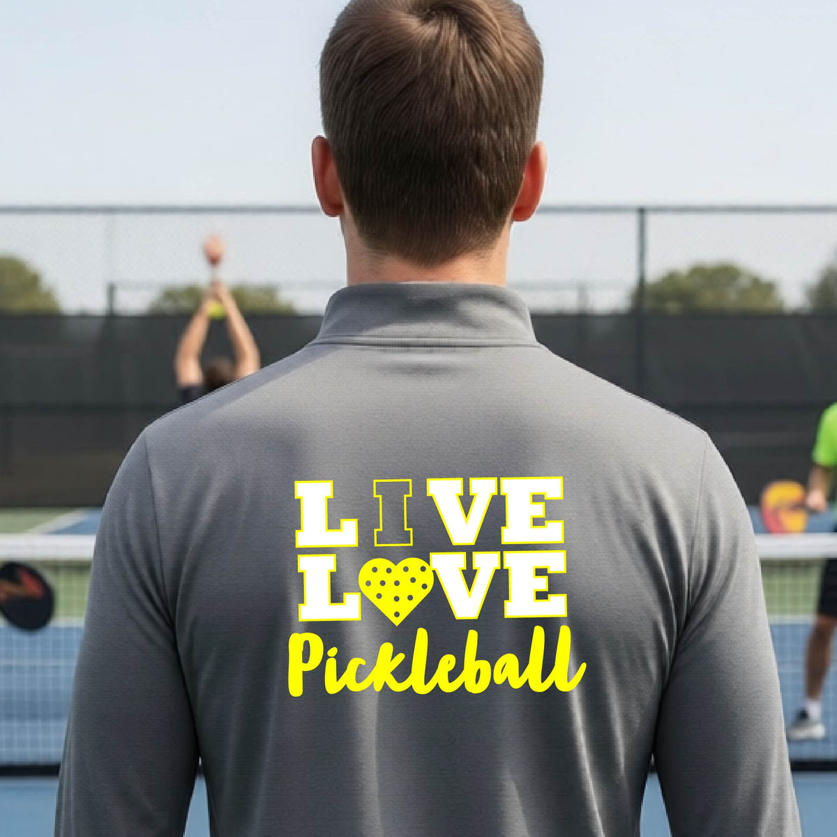 A man viewed from behind wearing a gray long-sleeve athletic quarter-zip shirt with a yellow graphic on the back that reads 'LIVE LOVE Pickleball'  with a pickleball motiff heart for replacing the O,set against an outdoor blurred background.