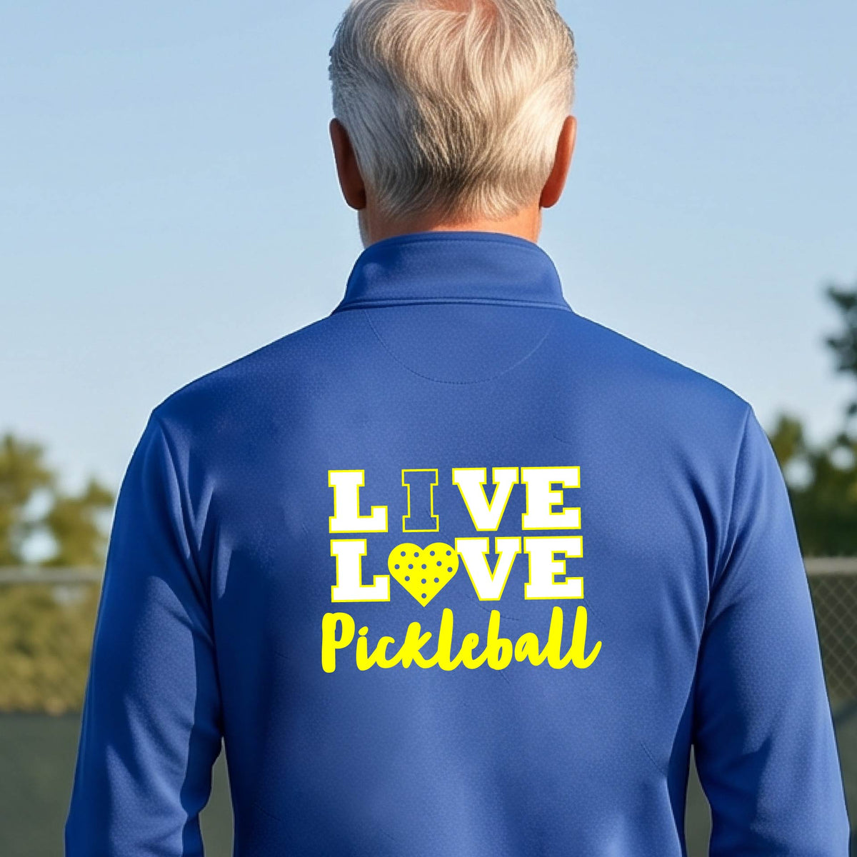 A man viewed from behind wearing a royal blue long-sleeve athletic quarter-zip shirt with a yellow graphic on the back that reads 'LIVE LOVE Pickleball'  with a pickleball motiff heart for replacing the O,set against an outdoor blurred background.