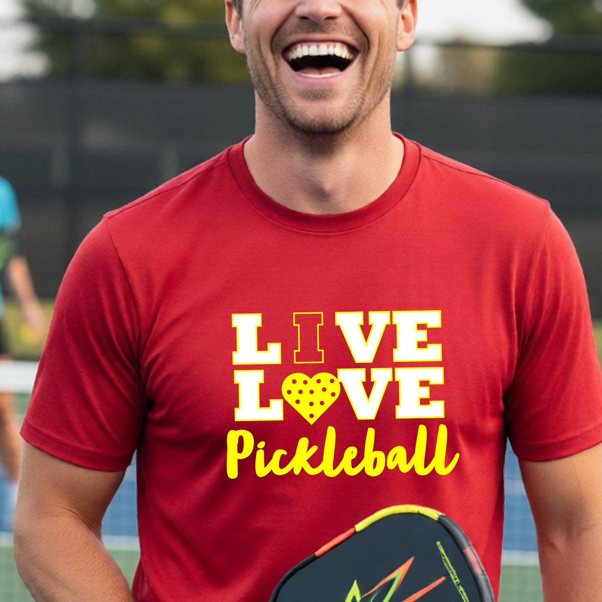 A man on a pickleball court wearing a red short-sleeve V-neck shirt with a yellow graphic that reads 'LIVE LOVE Pickleball' and a heart-shaped pickleball dot pattern inside the word 'LOVE'.