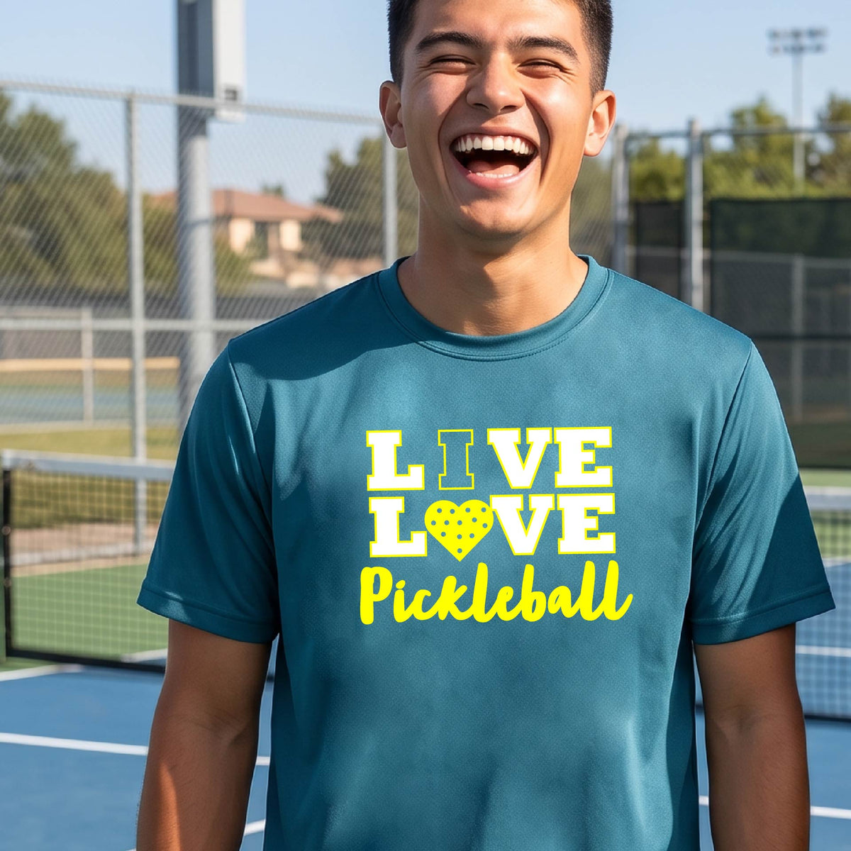 A man on a pickleball court wearing a teal blue short-sleeve V-neck shirt with a yellow graphic that reads 'LIVE LOVE Pickleball' and a heart-shaped pickleball dot pattern inside the word 'LOVE'.