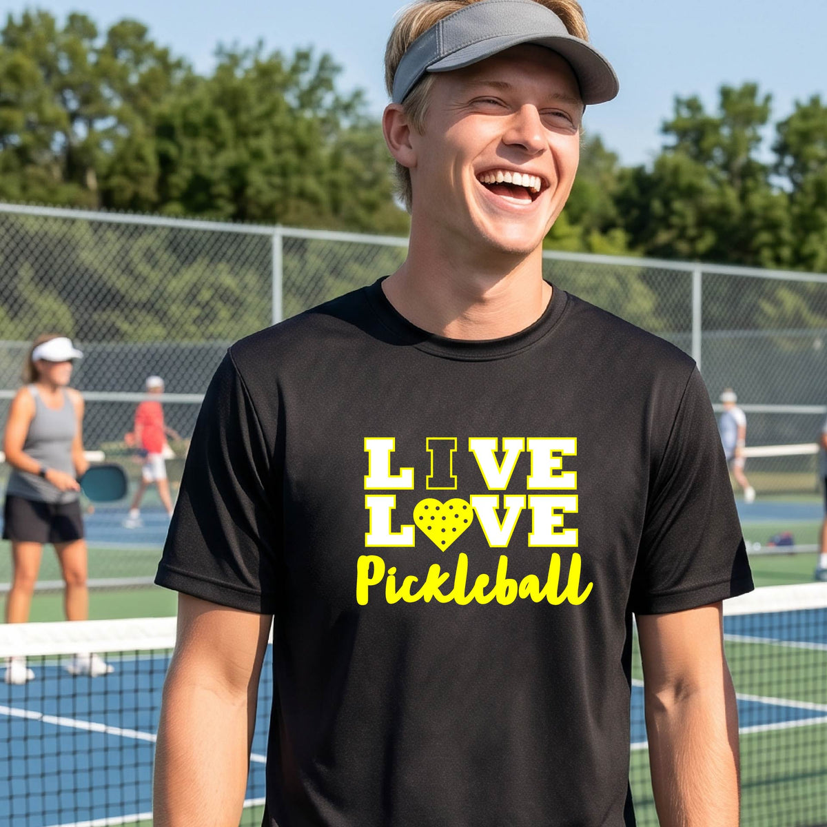 A man on a pickleball court wearing a black short-sleeve V-neck shirt with a yellow graphic that reads 'LIVE LOVE Pickleball' and a heart-shaped pickleball dot pattern inside the word 'LOVE'.
