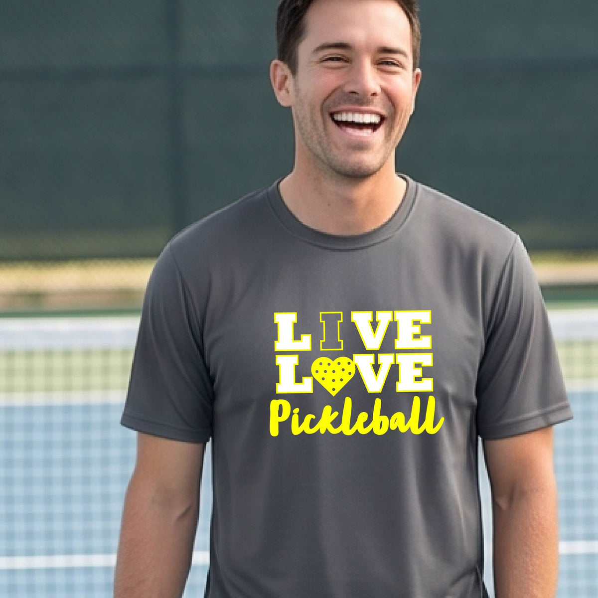 A man on a pickleball court wearing a dark gray short-sleeve V-neck shirt with a yellow graphic that reads 'LIVE LOVE Pickleball' and a heart-shaped pickleball dot pattern inside the word 'LOVE'.