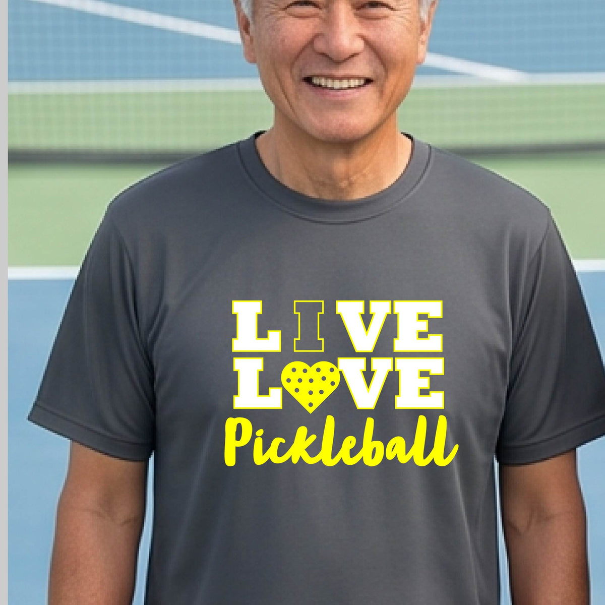 A man on a pickleball court wearing a dark gray short-sleeve V-neck shirt with a yellow graphic that reads 'LIVE LOVE Pickleball' and a heart-shaped pickleball dot pattern inside the word 'LOVE'.