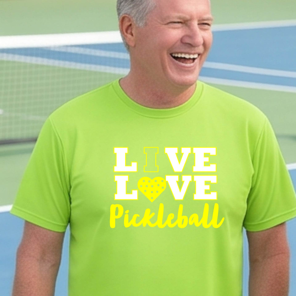 A man on a pickleball court wearing a lime green short-sleeve V-neck shirt with a yellow graphic that reads 'LIVE LOVE Pickleball' and a heart-shaped pickleball dot pattern inside the word 'LOVE'.