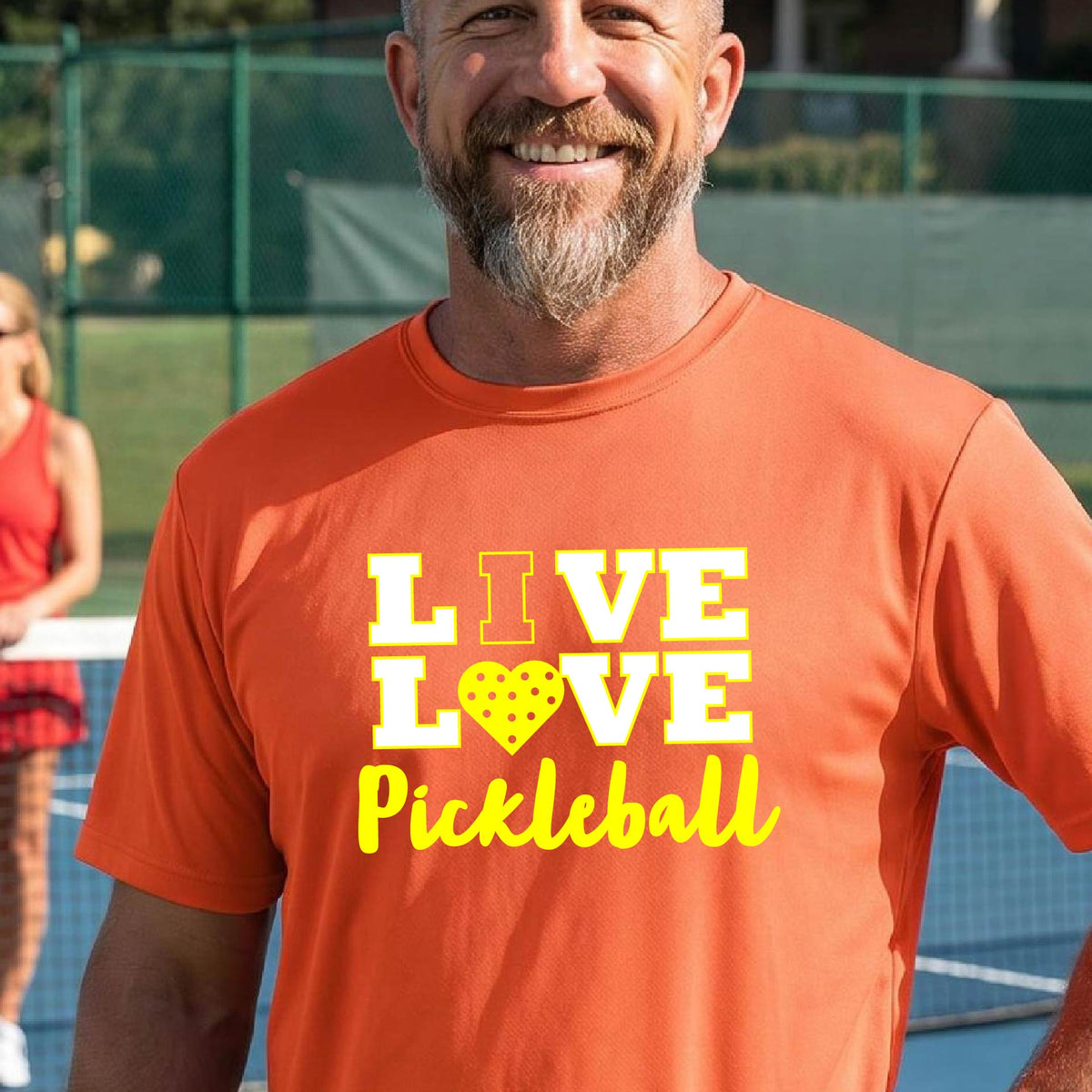 A man on a pickleball court wearing an orange short-sleeve V-neck shirt with a yellow graphic that reads 'LIVE LOVE Pickleball' and a heart-shaped pickleball dot pattern inside the word 'LOVE'.