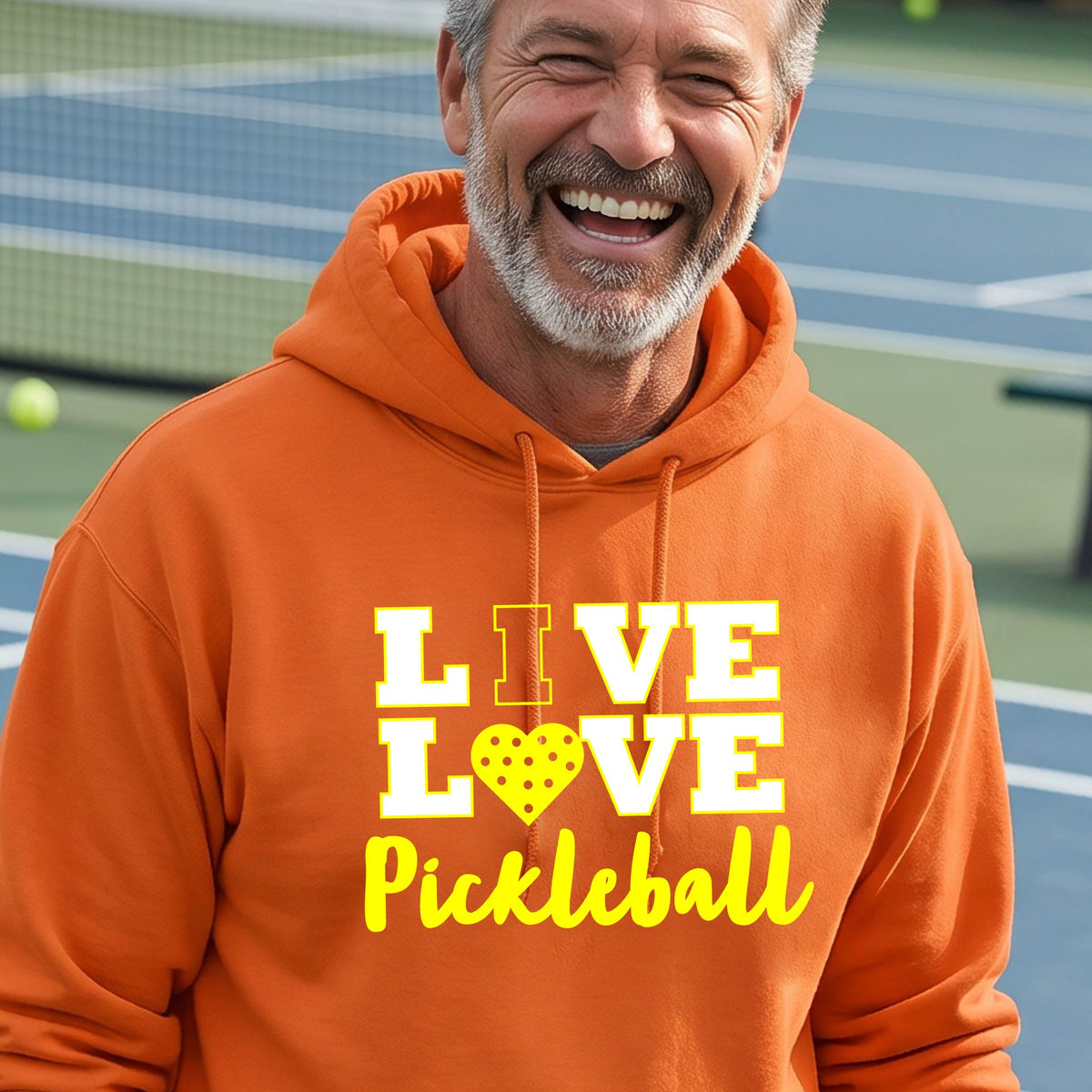 A man on a pickleball court wearing an orange unisex hoodie pullover shirt with a yellow graphic that reads 'LIVE LOVE Pickleball' and a heart-shaped pickleball dot pattern inside the word 'LOVE'