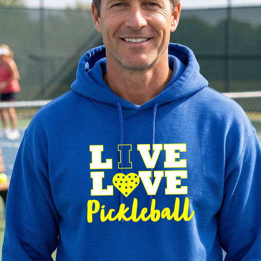 A man on a pickleball court wearing a royal blue unisex hoodie pullover shirt with a yellow graphic that reads 'LIVE LOVE Pickleball' and a heart-shaped pickleball dot pattern inside the word 'LOVE'