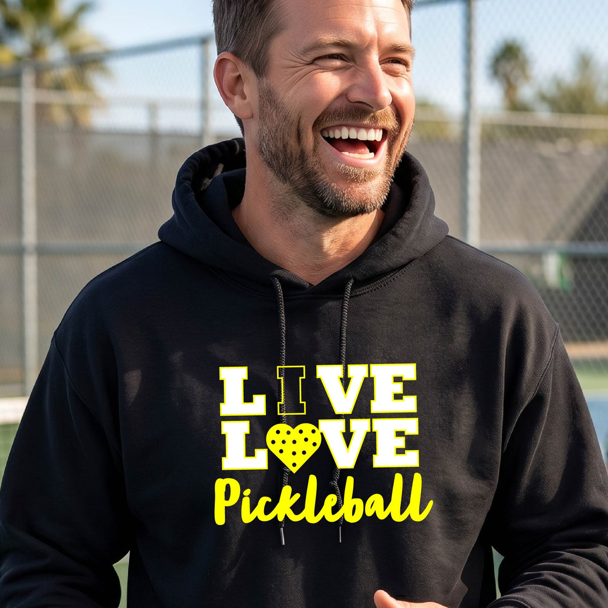 A man on a pickleball court wearing a black unisex hoodie pullover shirt with a yellow graphic that reads 'LIVE LOVE Pickleball' and a heart-shaped pickleball dot pattern inside the word 'LOVE'