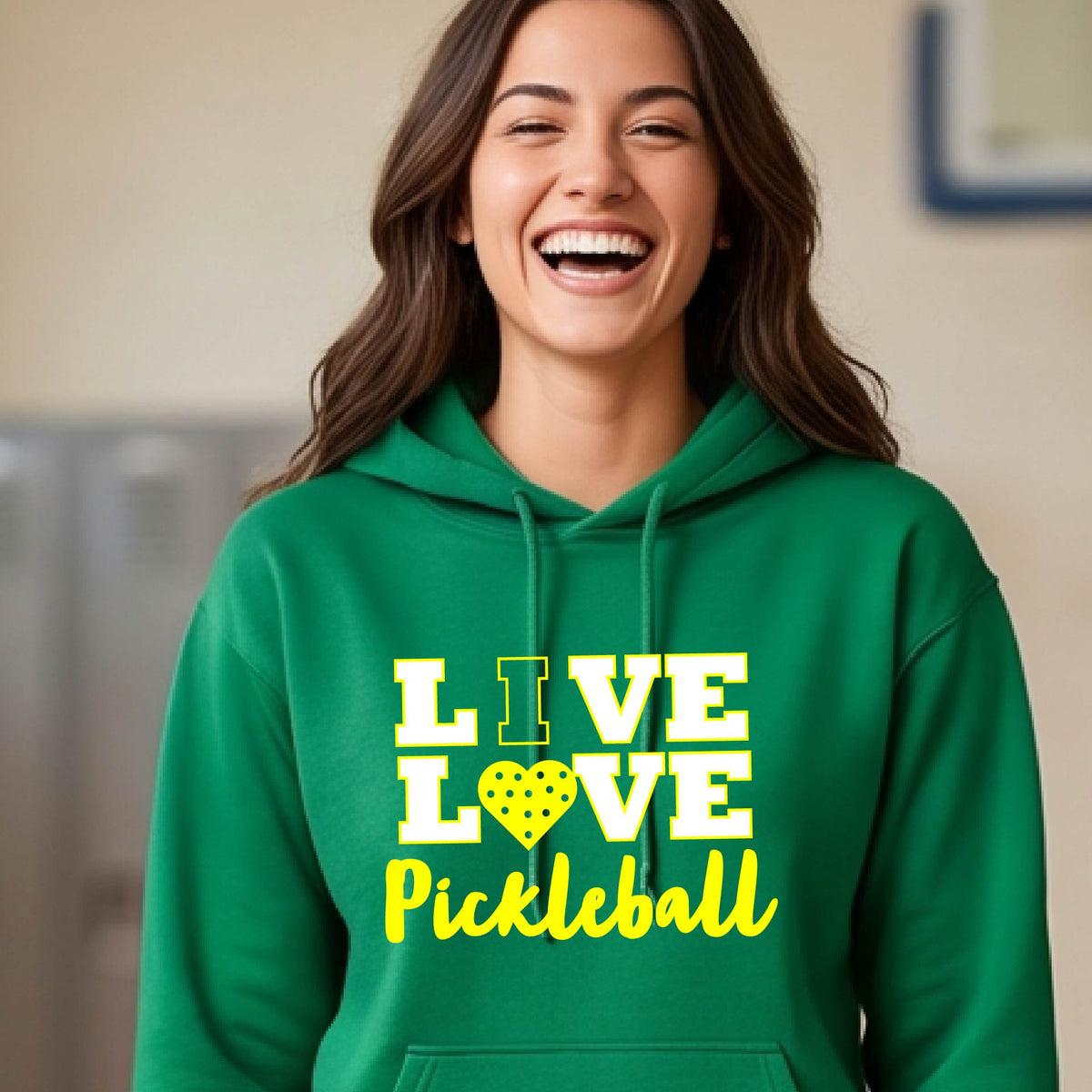 A woman on a pickleball court wearing a green unisex hoodie pullover shirt with a yellow graphic that reads 'LIVE LOVE Pickleball' and a heart-shaped pickleball dot pattern inside the word 'LOVE'