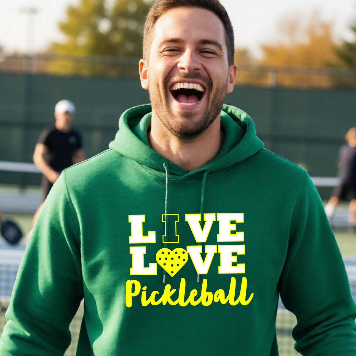 A man on a pickleball court wearing a green unisex hoodie pullover shirt with a yellow graphic that reads 'LIVE LOVE Pickleball' and a heart-shaped pickleball dot pattern inside the word 'LOVE'