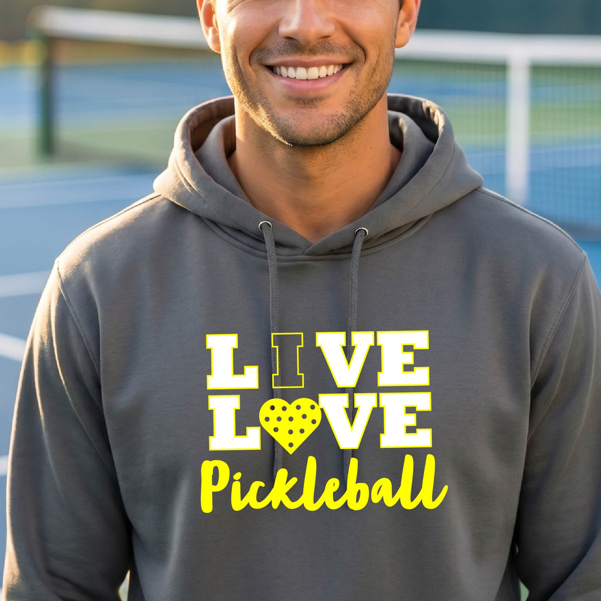 A man on a pickleball court wearing a gray unisex hoodie pullover shirt with a yellow graphic that reads 'LIVE LOVE Pickleball' and a heart-shaped pickleball dot pattern inside the word 'LOVE'