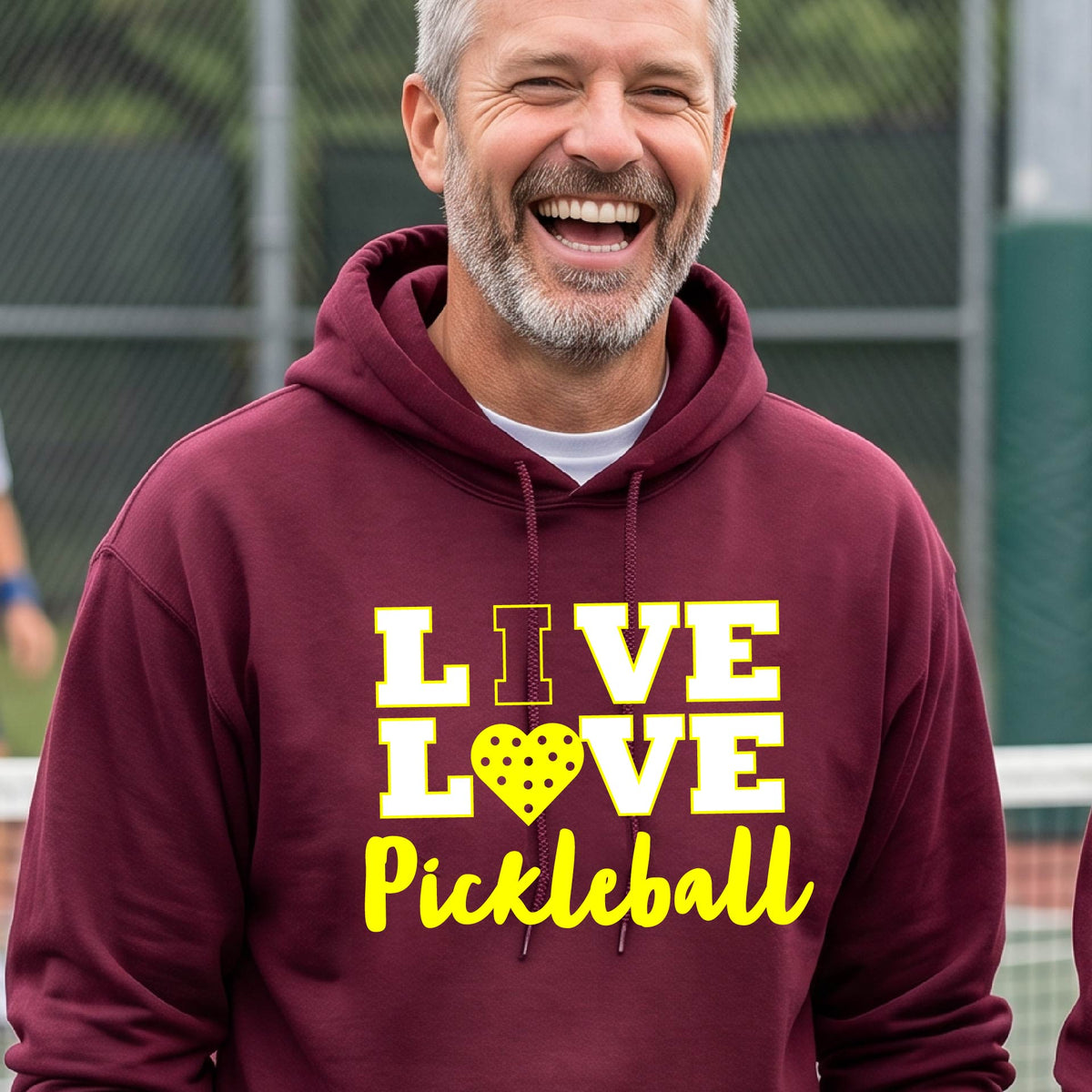 A man on a pickleball court wearing a maroon unisex hoodie pullover shirt with a yellow graphic that reads 'LIVE LOVE Pickleball' and a heart-shaped pickleball dot pattern inside the word 'LOVE'