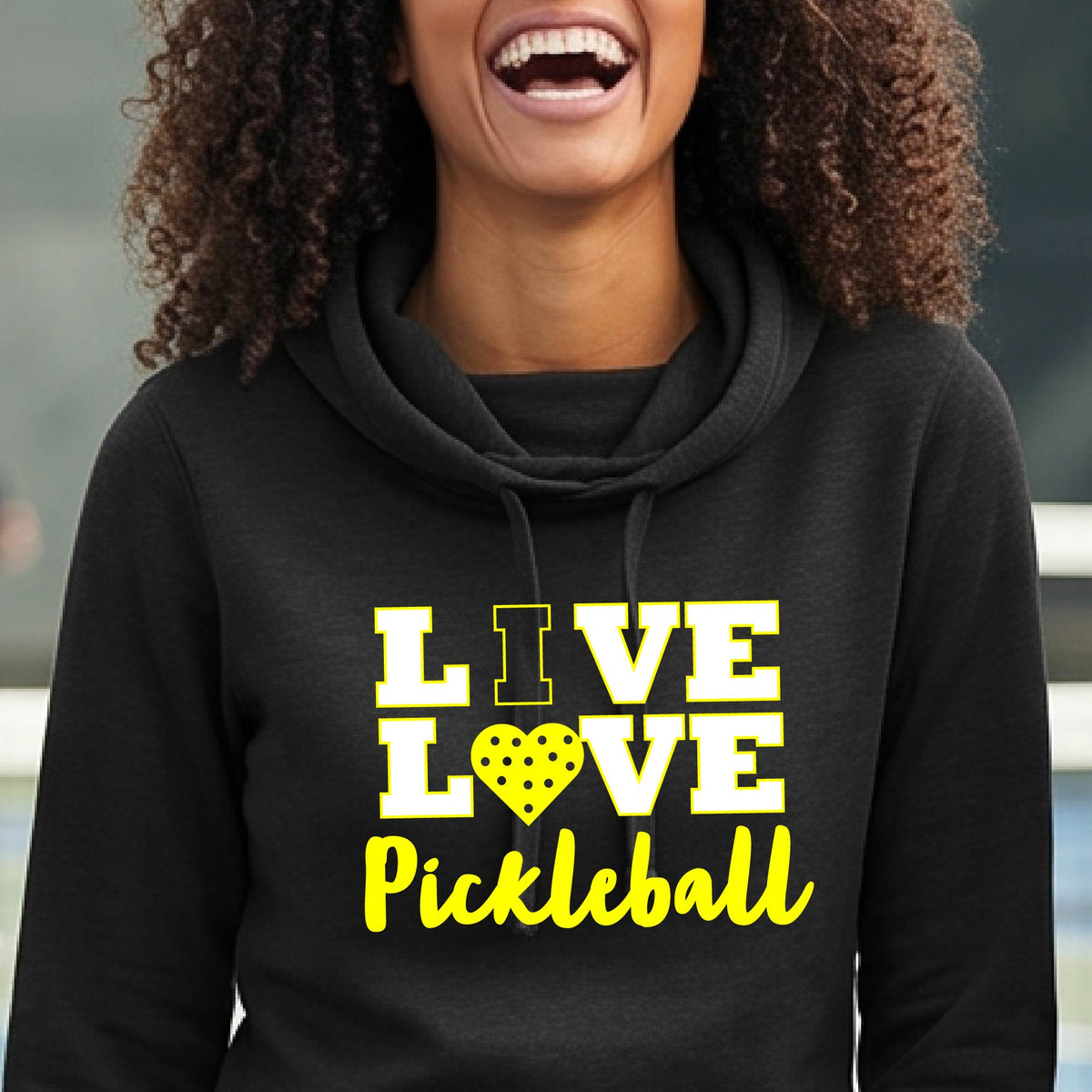 A smiling woman wearing a black pullover hoodie with a yellow graphic that reads 'LIVE LOVE Pickleball' and a pickleball icon, photographed outdoors with hills and a fence in the background.