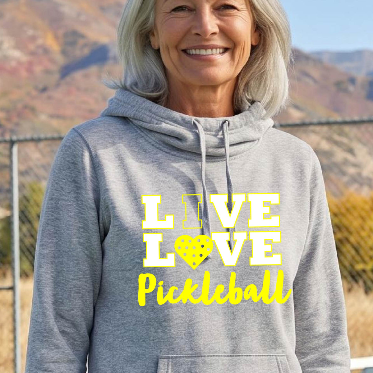 A smiling older woman wearing a heather gray pullover hoodie with a yellow graphic that reads 'LIVE LOVE Pickleball' and a pickleball icon, photographed outdoors with hills and a fence in the background.