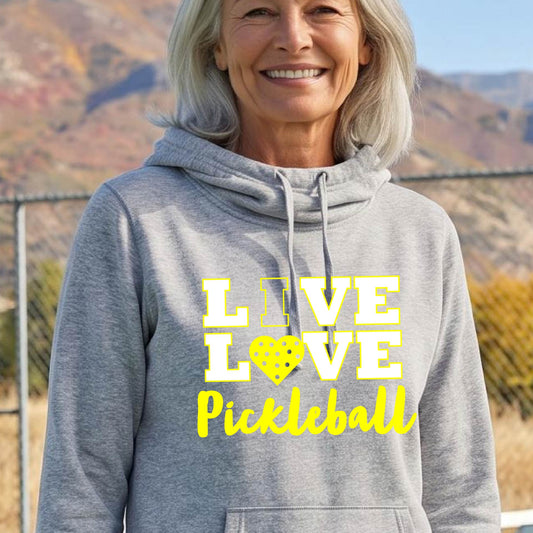 A smiling older woman wearing a heather gray pullover hoodie with a yellow graphic that reads 'LIVE LOVE Pickleball' and a pickleball icon, photographed outdoors with hills and a fence in the background.