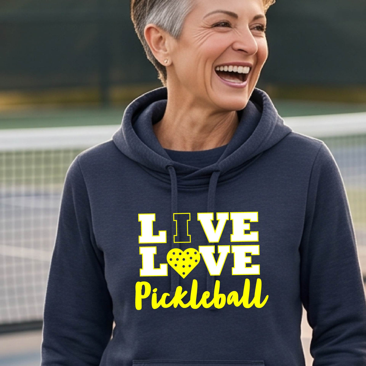 A smiling older woman wearing anavy blue pullover hoodie with a yellow graphic that reads 'LIVE LOVE Pickleball' and a pickleball icon, photographed outdoors with hills and a fence in the background.