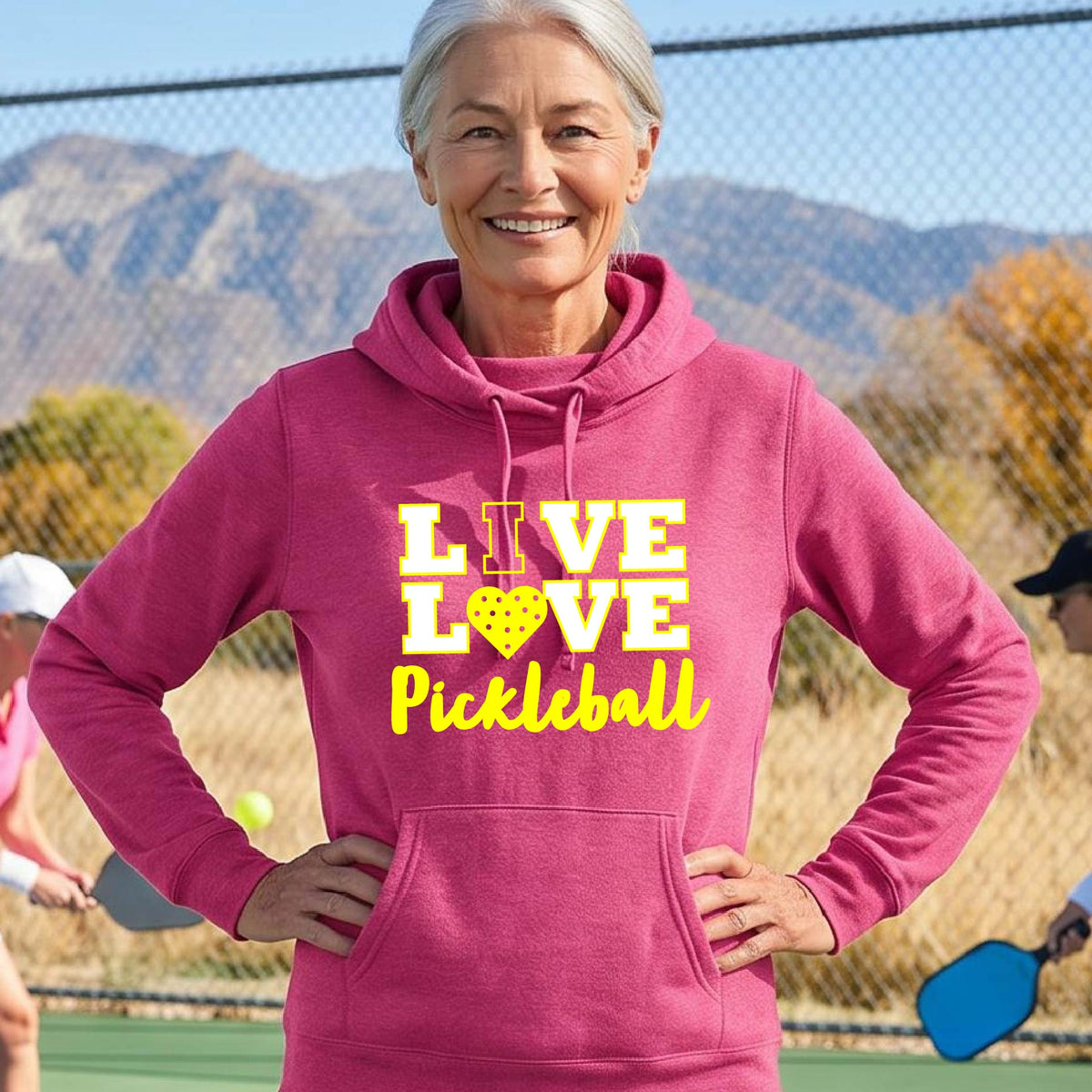 A smiling older woman wearing a pink pullover hoodie with a yellow graphic that reads 'LIVE LOVE Pickleball' and a pickleball icon, photographed outdoors with hills and a fence in the background.