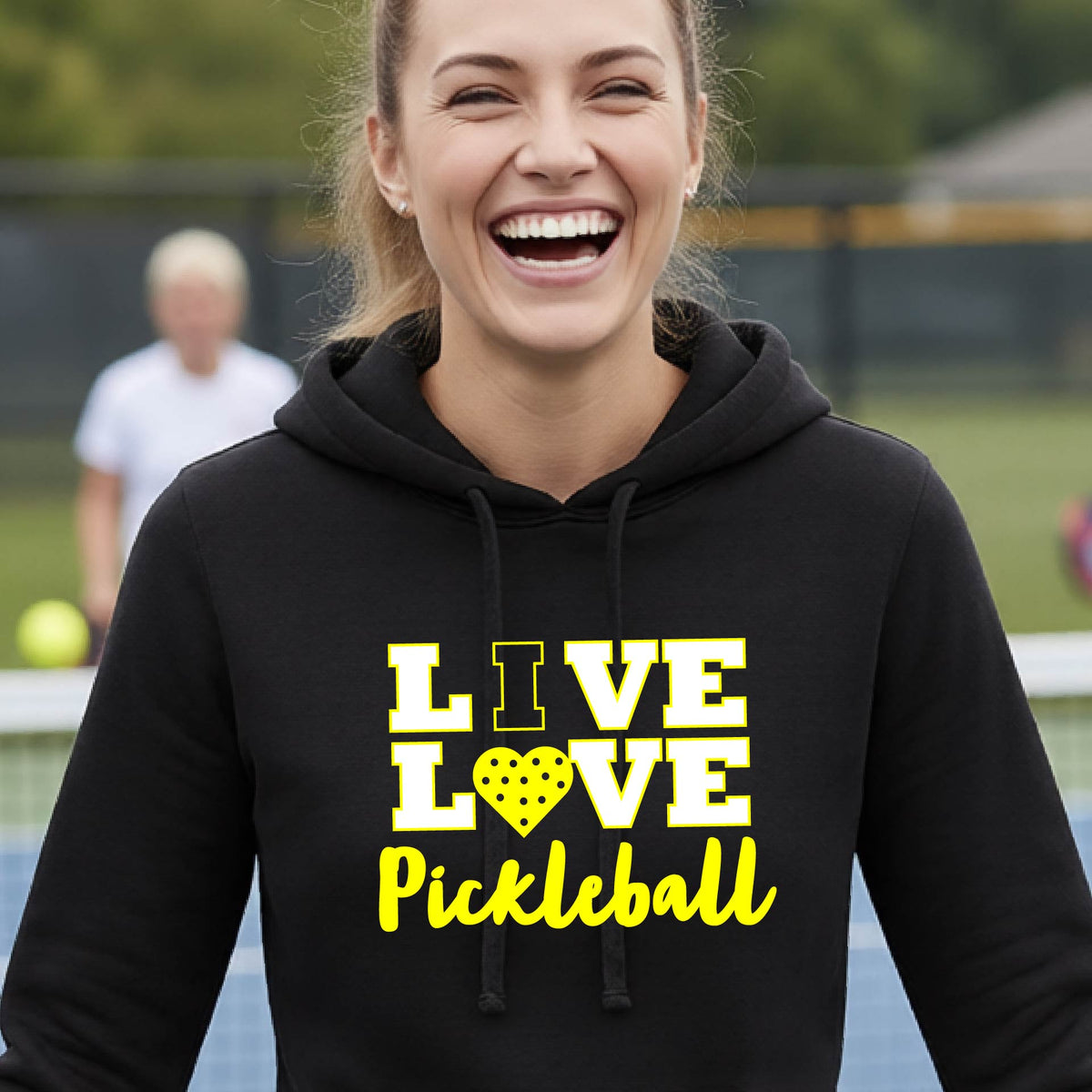 A woman on a pickleball court wearing a black fitted hoodie pullover sweatshirt with a yellow graphic that reads 'LIVE LOVE Pickleball' and a heart-shaped pickleball dot pattern inside the word 'LOVE'