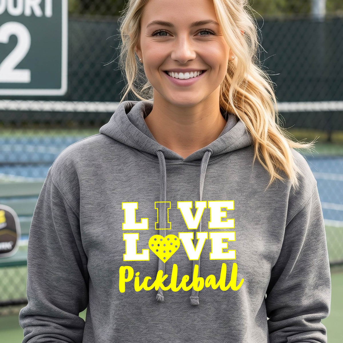A smiling blonde woman wearing a heather gray pullover hoodie with a yellow graphic that reads 'I LOVE PICKLEBALL' with a heart-shaped pickleball motif, photographed on a pickleball court with a chain-link fence in the background.