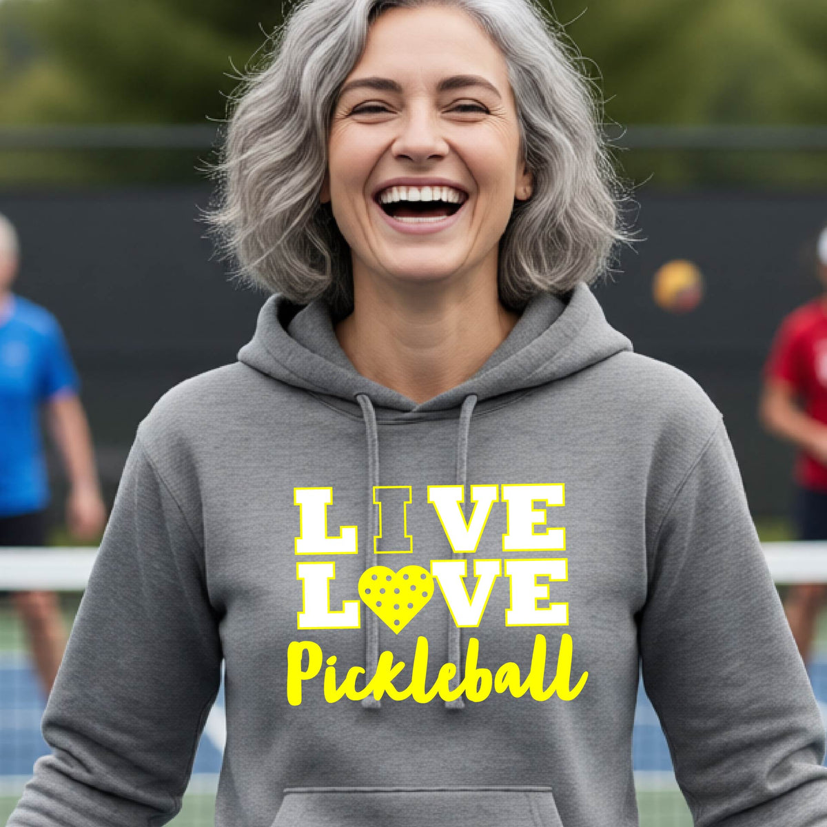 A woman on a pickleball court wearing a light gray fitted hoodie pullover sweatshirt with a yellow graphic that reads 'LIVE LOVE Pickleball' and a heart-shaped pickleball dot pattern inside the word 'LOVE'