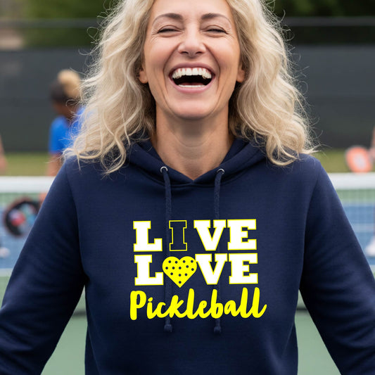 A woman on a pickleball court wearing a navy blue fitted hoodie pullover sweatshirt with a yellow graphic that reads 'LIVE LOVE Pickleball' and a heart-shaped pickleball dot pattern inside the word 'LOVE'