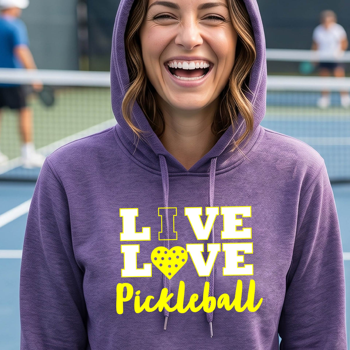 A woman on a pickleball court wearing a purple fitted hoodie pullover sweatshirt with a yellow graphic that reads 'LIVE LOVE Pickleball' and a heart-shaped pickleball dot pattern inside the word 'LOVE'
