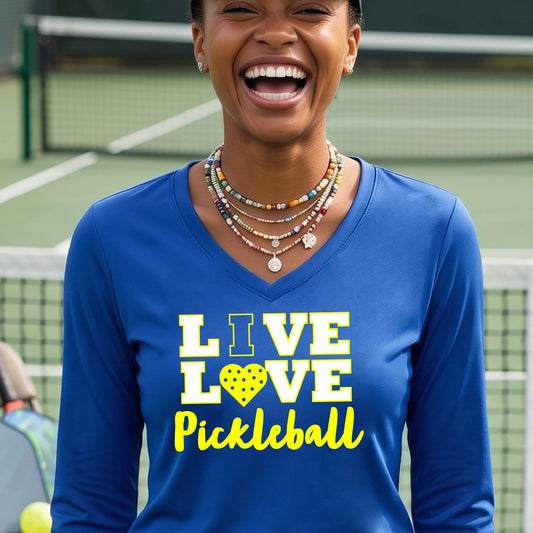 A woman on a pickleball court wearing a royal blue long-sleeve V-neck shirt with a yellow graphic that reads 'LIVE LOVE Pickleball' and a heart-shaped pickleball dot pattern inside the word 'LOVE'.