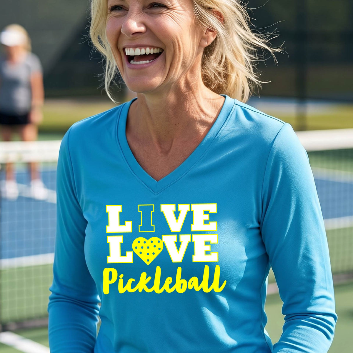 A woman on a pickleball court wearing a bright blue long-sleeve V-neck shirt with a yellow graphic that reads 'LIVE LOVE Pickleball' and a heart-shaped pickleball dot pattern inside the word 'LOVE'.