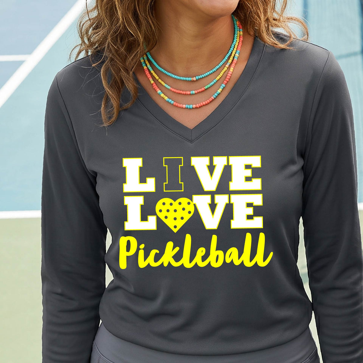 A woman on a pickleball court wearing a dark gray long-sleeve V-neck shirt with a yellow graphic that reads 'LIVE LOVE Pickleball' and a heart-shaped pickleball dot pattern inside the word 'LOVE'.