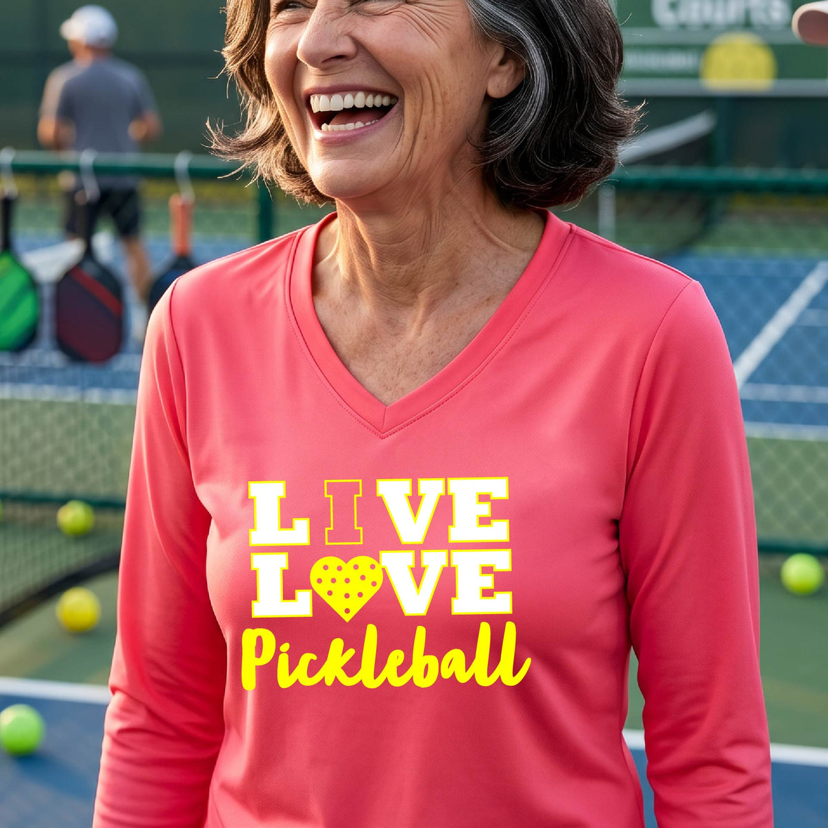 A woman on a pickleball court wearing a coral pink long-sleeve V-neck shirt with a yellow graphic that reads 'LIVE LOVE Pickleball' and a heart-shaped pickleball dot pattern inside the word 'LOVE'.