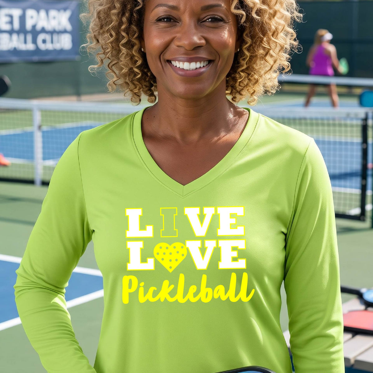 A woman on a pickleball court wearing a lime green long-sleeve V-neck shirt with a yellow graphic that reads 'LIVE LOVE Pickleball' and a heart-shaped pickleball dot pattern inside the word 'LOVE'.