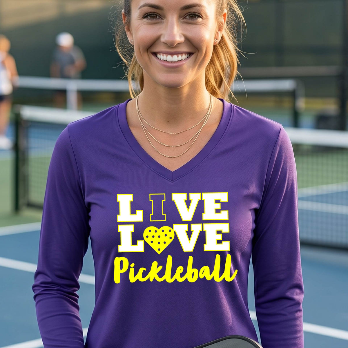 A woman on a pickleball court wearing a  purple long-sleeve V-neck shirt with a yellow graphic that reads 'LIVE LOVE Pickleball' and a heart-shaped pickleball dot pattern inside the word 'LOVE'. 