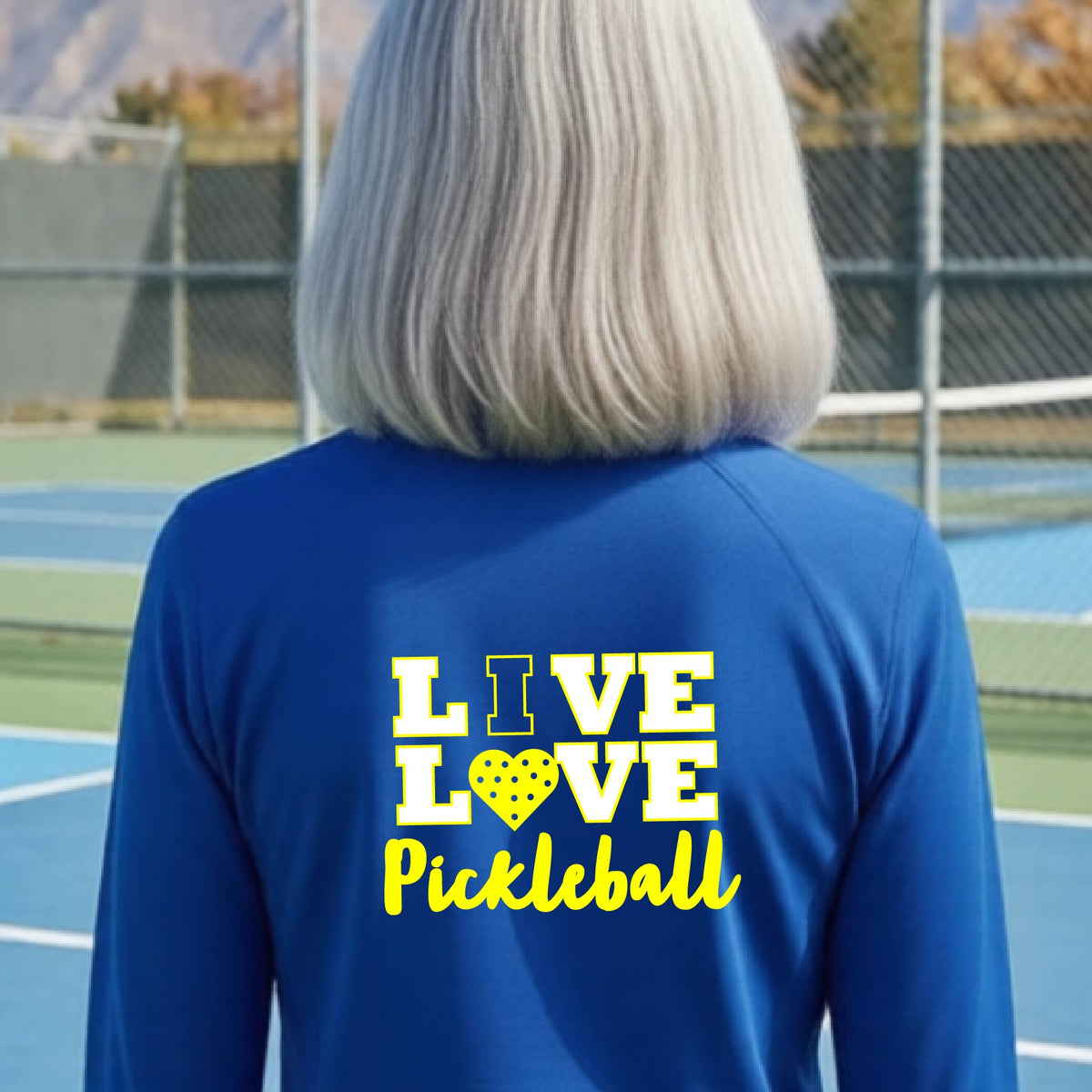 A woman viewed from behind wearing a bright royal blue quarter-zip athletic pullover with a yellow and white graphic on the back that reads 'LIVE LOVE Pickleball'.