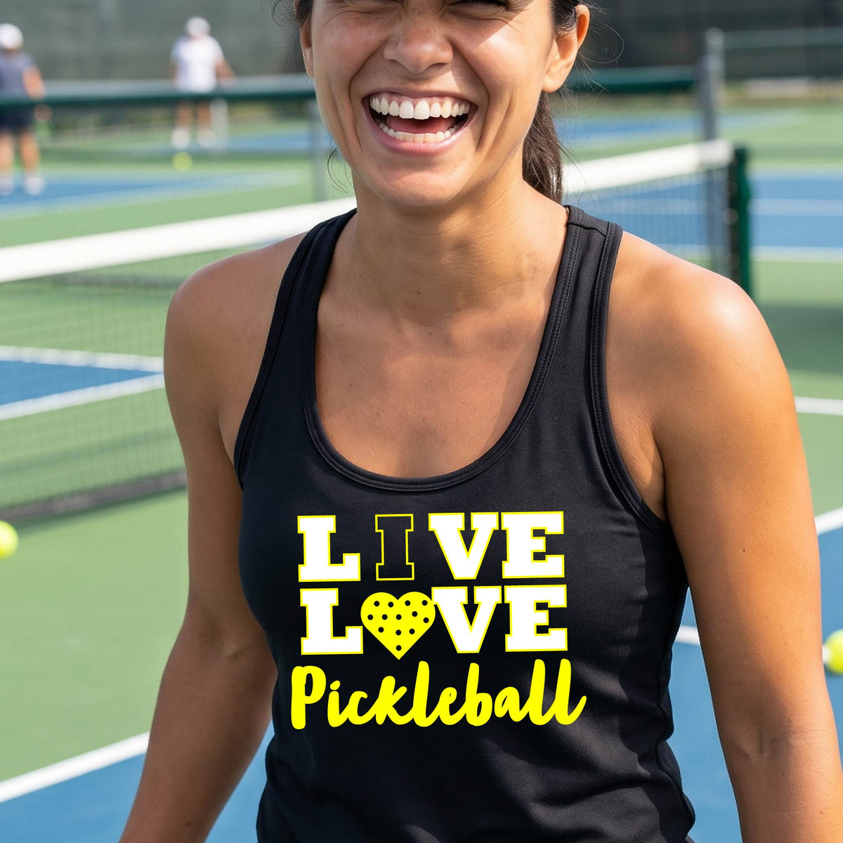A woman on a pickleball court wearing a black tank top with a yellow and white graphic that reads 'LIVE LOVE Pickleball' with a heart shaped pickleball motiff replace the O.