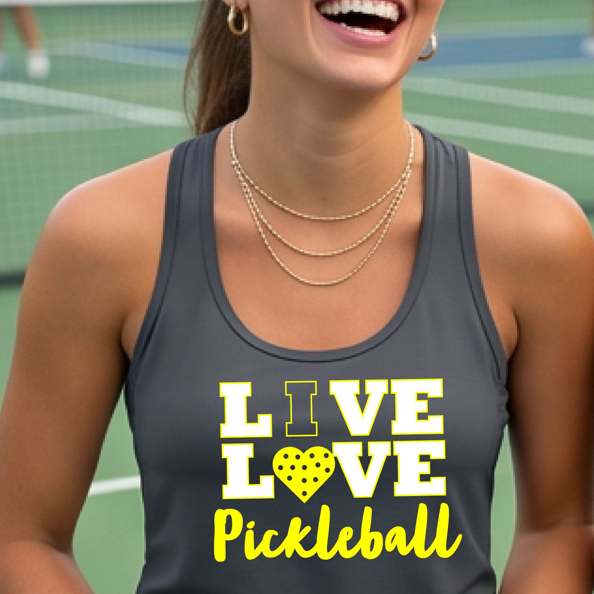 A woman on a pickleball court wearing a dark gray tank top with a yellow and white graphic that reads 'LIVE LOVE Pickleball' with a heart shaped pickleball motiff replace the O.