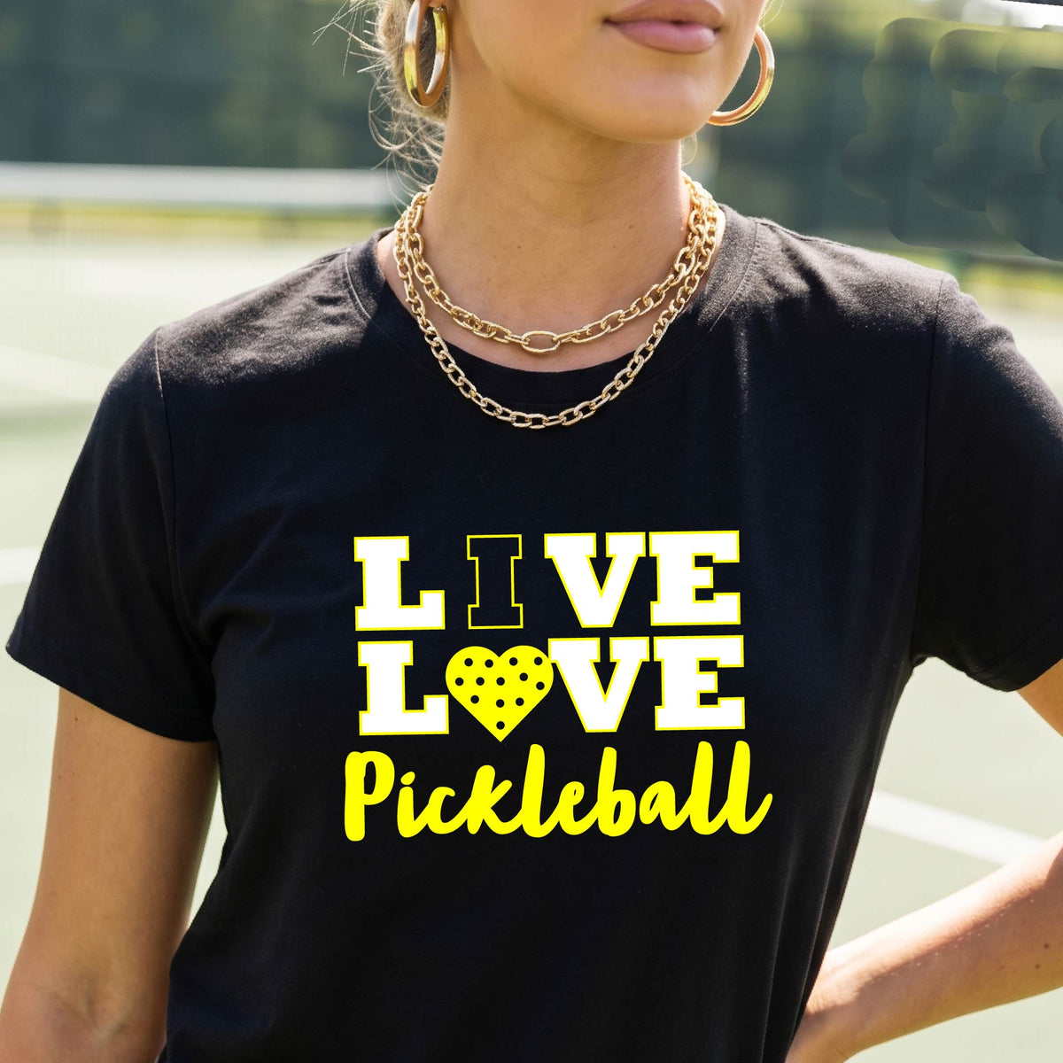 A woman on a pickleball court wearing a black short-sleeve t-shirt with a white and yellow graphic that reads 'LIVE LOVE Pickleball' and a heart-shaped pickleball dot pattern inside the word 'LOVE'.