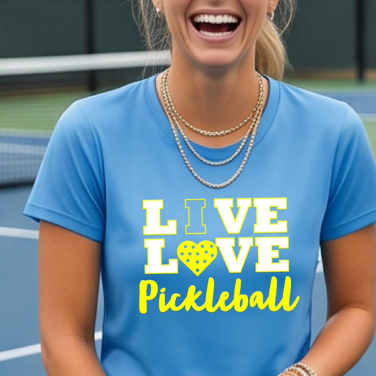 A woman on a pickleball court wearing a bright blue short-sleeve t-shirt with a white and yellow graphic that reads 'LIVE LOVE Pickleball' and a heart-shaped pickleball dot pattern inside the word 'LOVE'.
