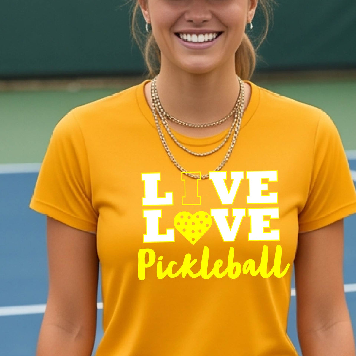A woman on a pickleball court wearing a mustard yellow gold short-sleeve t-shirt with a white and yellow graphic that reads 'LIVE LOVE Pickleball' and a heart-shaped pickleball dot pattern inside the word 'LOVE'.