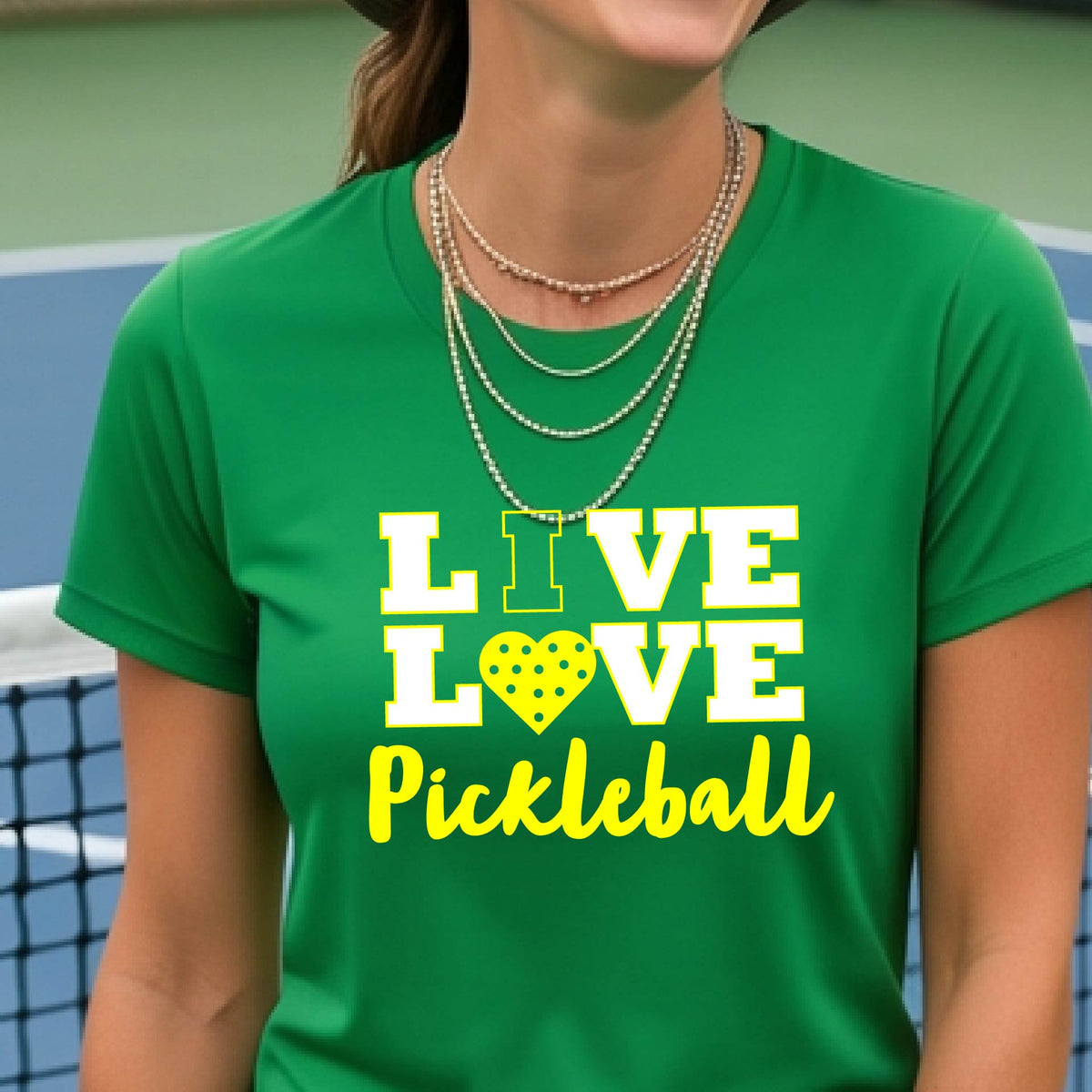 A woman on a pickleball court wearing a green short-sleeve t-shirt with a white and yellow graphic that reads 'LIVE LOVE Pickleball' and a heart-shaped pickleball dot pattern inside the word 'LOVE'.


