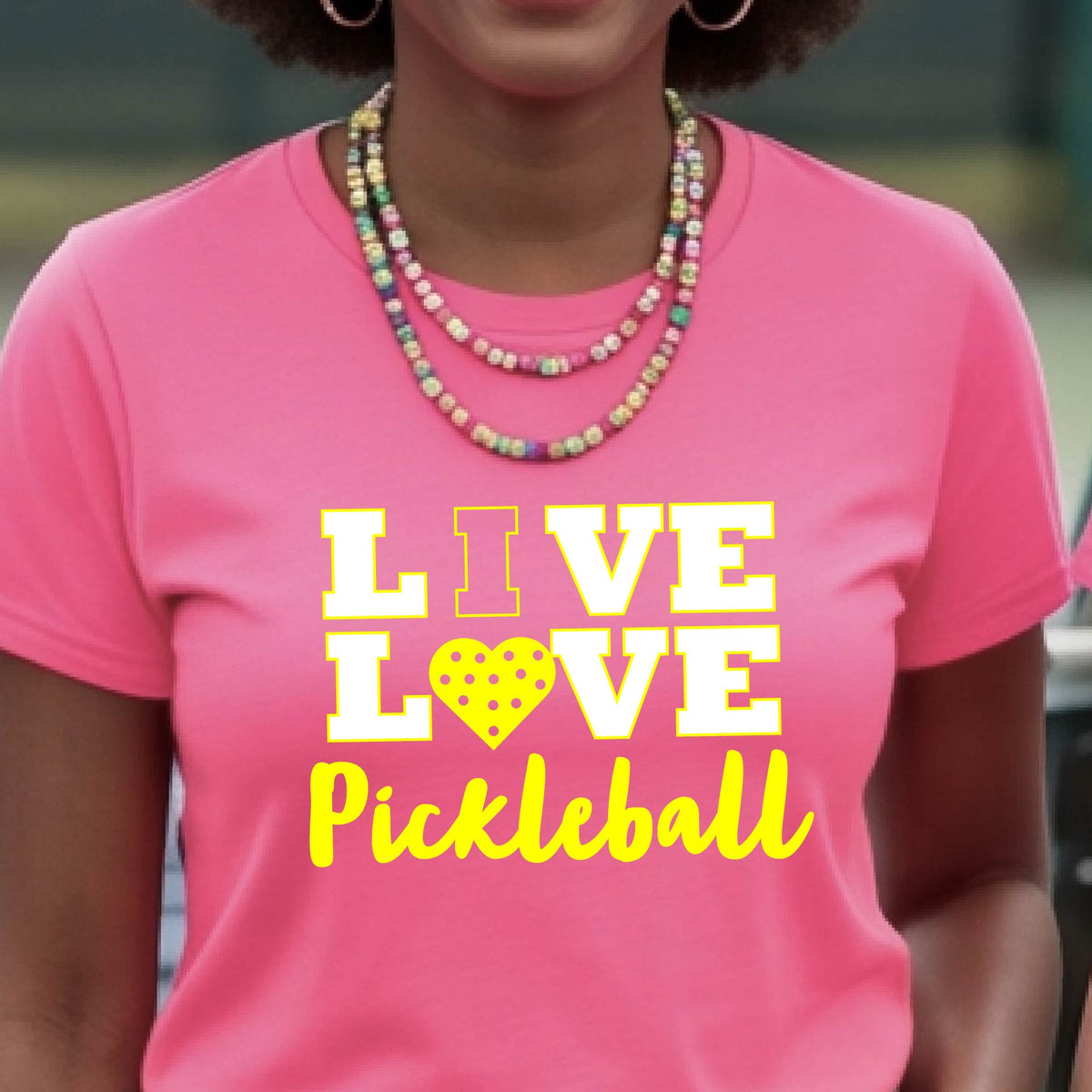 A woman on a pickleball court wearing a pink short-sleeve t-shirt with a white and yellow graphic that reads 'LIVE LOVE Pickleball' and a heart-shaped pickleball dot pattern inside the word 'LOVE'.